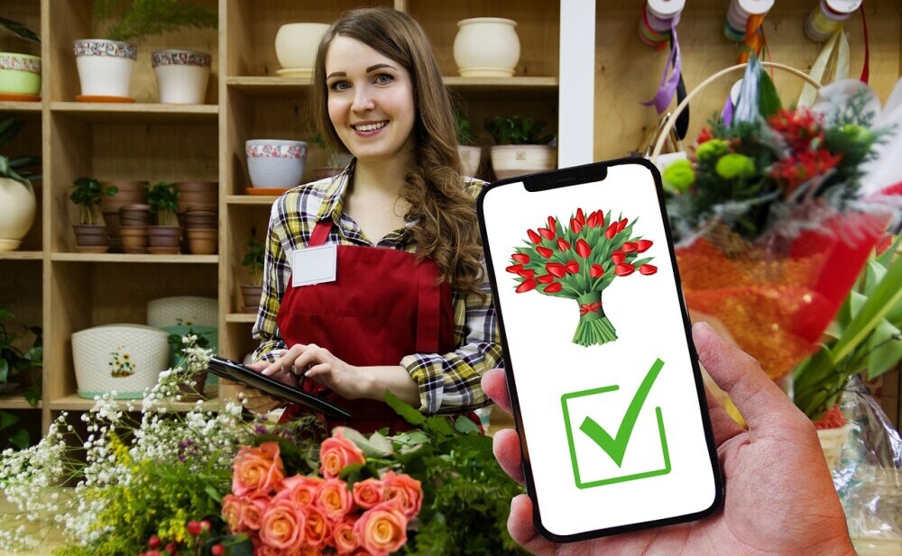 woman in flower shop smiling holding a tablet. flowers in front of her and a hand holding a phone with a picture of a flower with a tick sign below